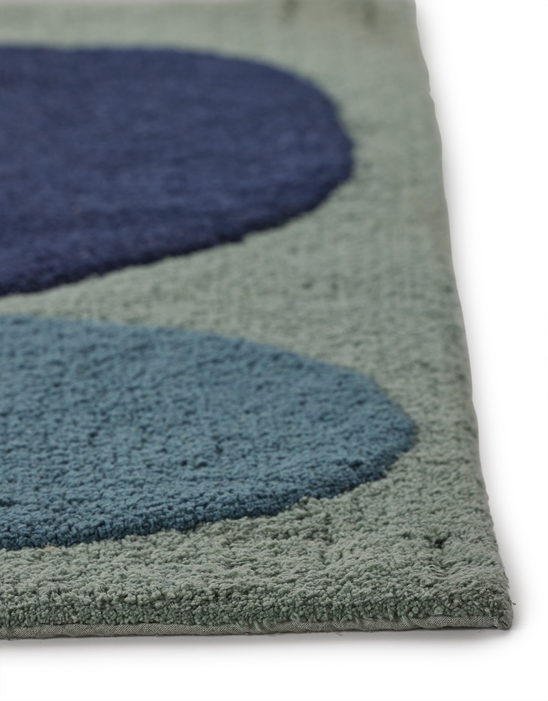 A close-up, angled view of a light teal angela adams "Two Stones Bluebird" bath mat. The image focuses on the texture of the pile and the raised, rounded shapes in navy blue and lighter blue, showing the distinct borders between the colors and the depth of the tufted material.