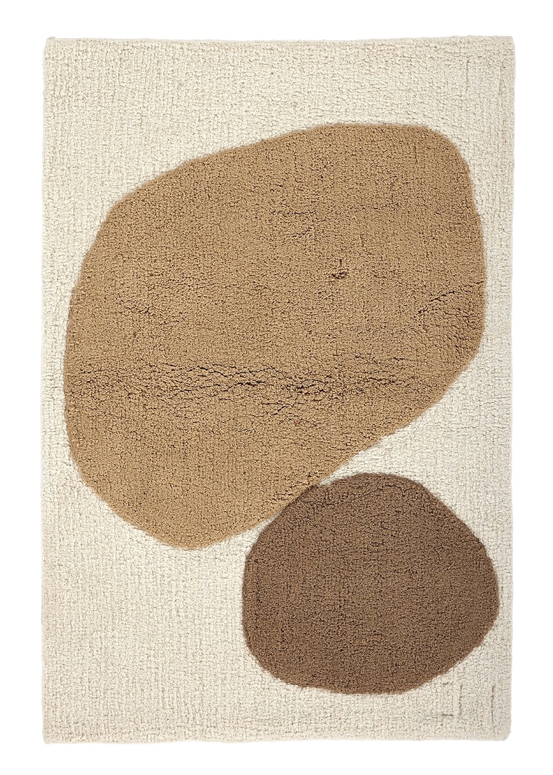 A light beige angela adams "Two Stones Sand" bath mat is viewed from directly above. It features two large, rounded, organic shapes: a lighter sandy brown one at the top and a smaller, darker brown one below it. The background of the mat is a soft, textured light beige.