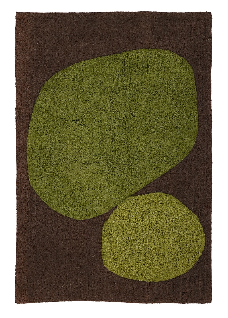 A brown and lime green abstract bath mat with two rounded, organic shapes is laid out flat on a vibrant green grassy lawn dotted with small white and yellow wildflowers. The mat is seen from directly above, with the larger lime green shape on the left and a smaller, slightly lighter lime green shape to its right, all against a dark brown background.