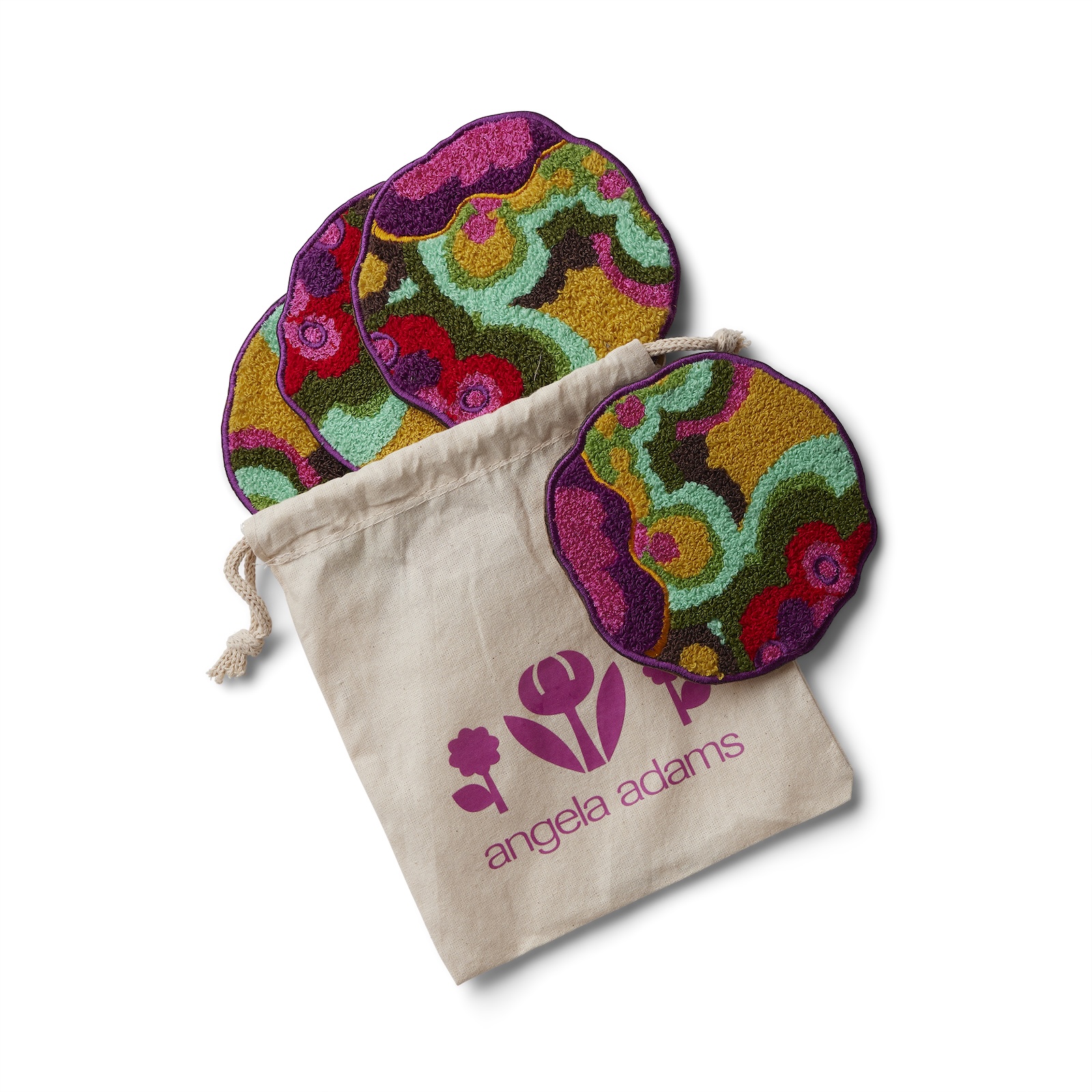 An overhead shot on a white background features a set of four round, irregularly shaped coasters with a vibrant, abstract floral pattern and a drawstring pouch. The coasters display shades of pink, fuchsia, purple, teal, yellow, and red. One coaster is leaning against the side of a light beige canvas pouch, while the other three are stacked behind it. The pouch has a fuchsia-colored logo depicting a flower with four petals and a stylized tulip, with the words "angela adams" written below it in a matching fuchsia font. The drawstring of the pouch is visible at the top left.