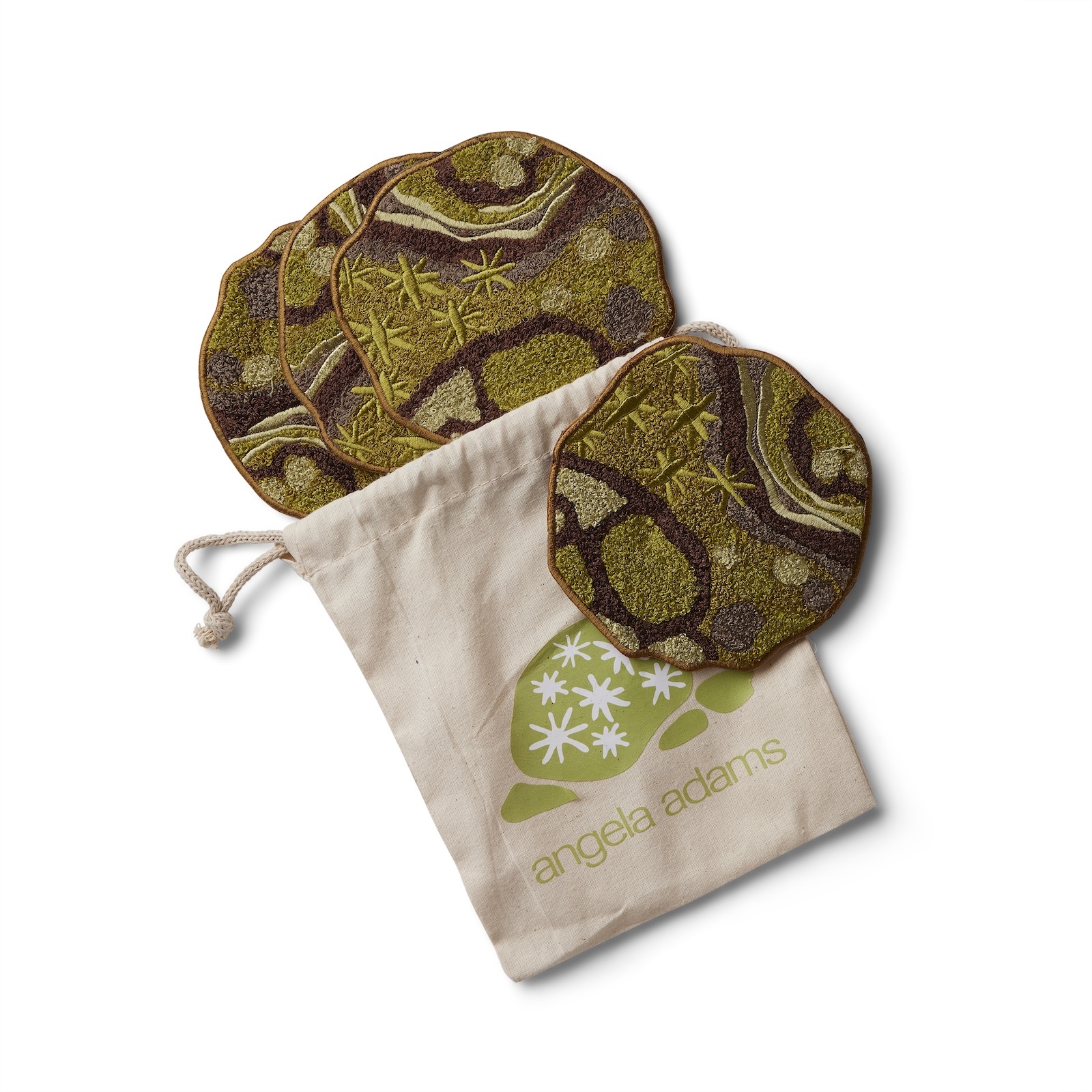 An overhead shot on a white background features a set of four round, irregularly shaped coasters with a brown, olive green, and light green abstract pattern, and a drawstring pouch. The pattern on the coasters resembles organic shapes and some have starburst-like designs. One coaster is leaning against the side of a light beige canvas pouch, while the other three are stacked behind it. The pouch has a lime green logo depicting a stylized leaf or organic shape with white starbursts, and the words "angela adams" written below it in a matching lime green font. The drawstring of the pouch is visible at the top left.