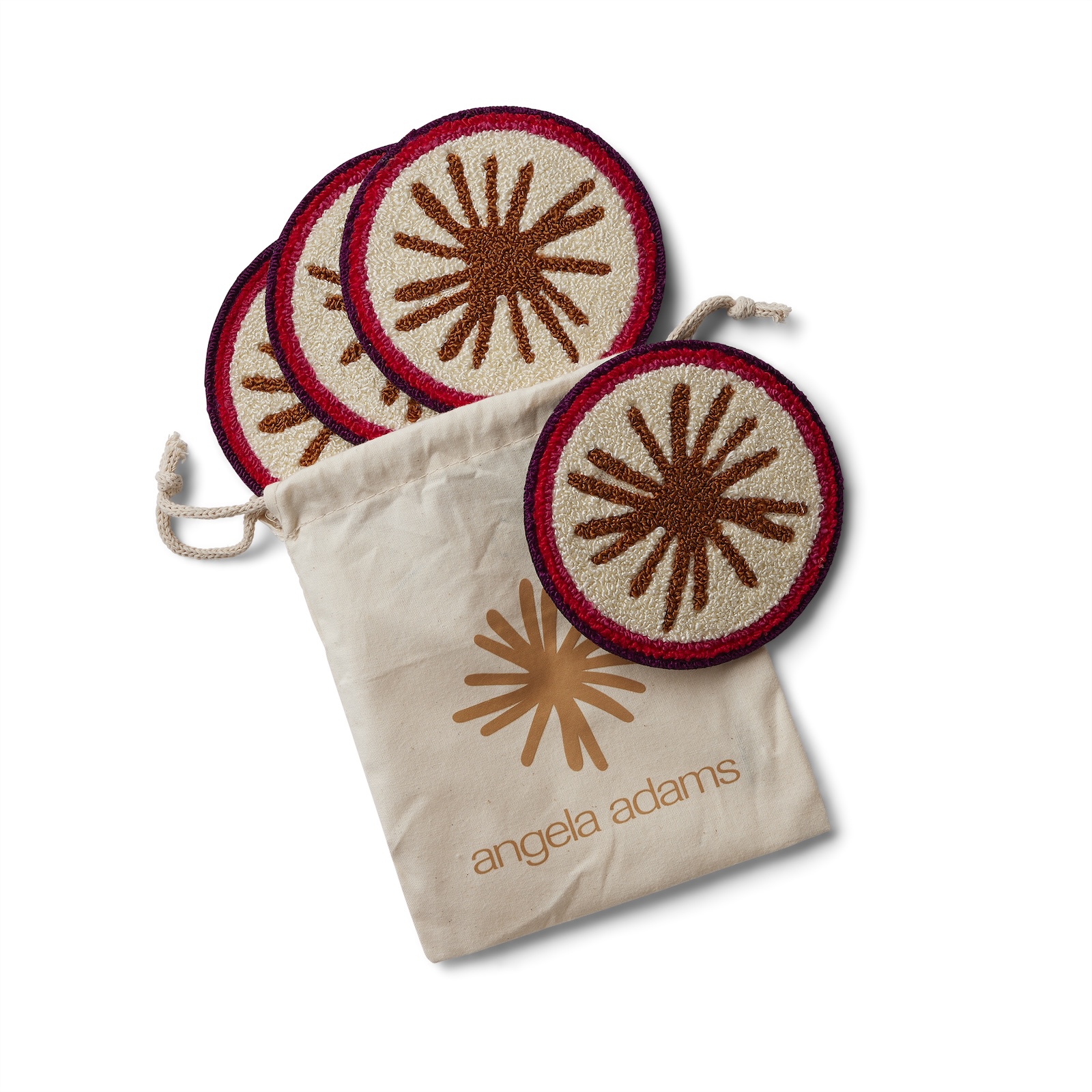 An overhead shot on a white background features a set of four round coasters and a drawstring pouch. The coasters have a cream-colored center with a brown starburst design and a red border. One coaster is leaning against the side of a light beige canvas pouch, while the other three are stacked behind it. The pouch has a faint, light brown starburst logo with the words "angela adams" written below it in a matching brown font. The drawstring of the pouch is visible at the top left.