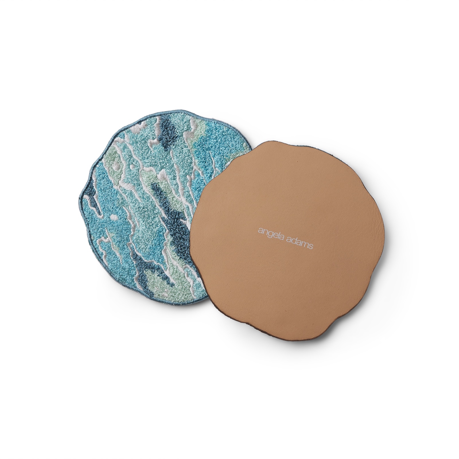 Two round coasters are shown side by side on a white background. One coaster displays a highly textured, tufted surface in oceanic hues of turquoise, teal, seafoam, and white, mimicking the movement of waves. The other coaster is turned over to reveal a smooth, tan leather backing with a soft, organic edge and the name “angela adams” printed in white at the center. The image highlights the contrast between the artistic front and the refined, durable back of the coaster.