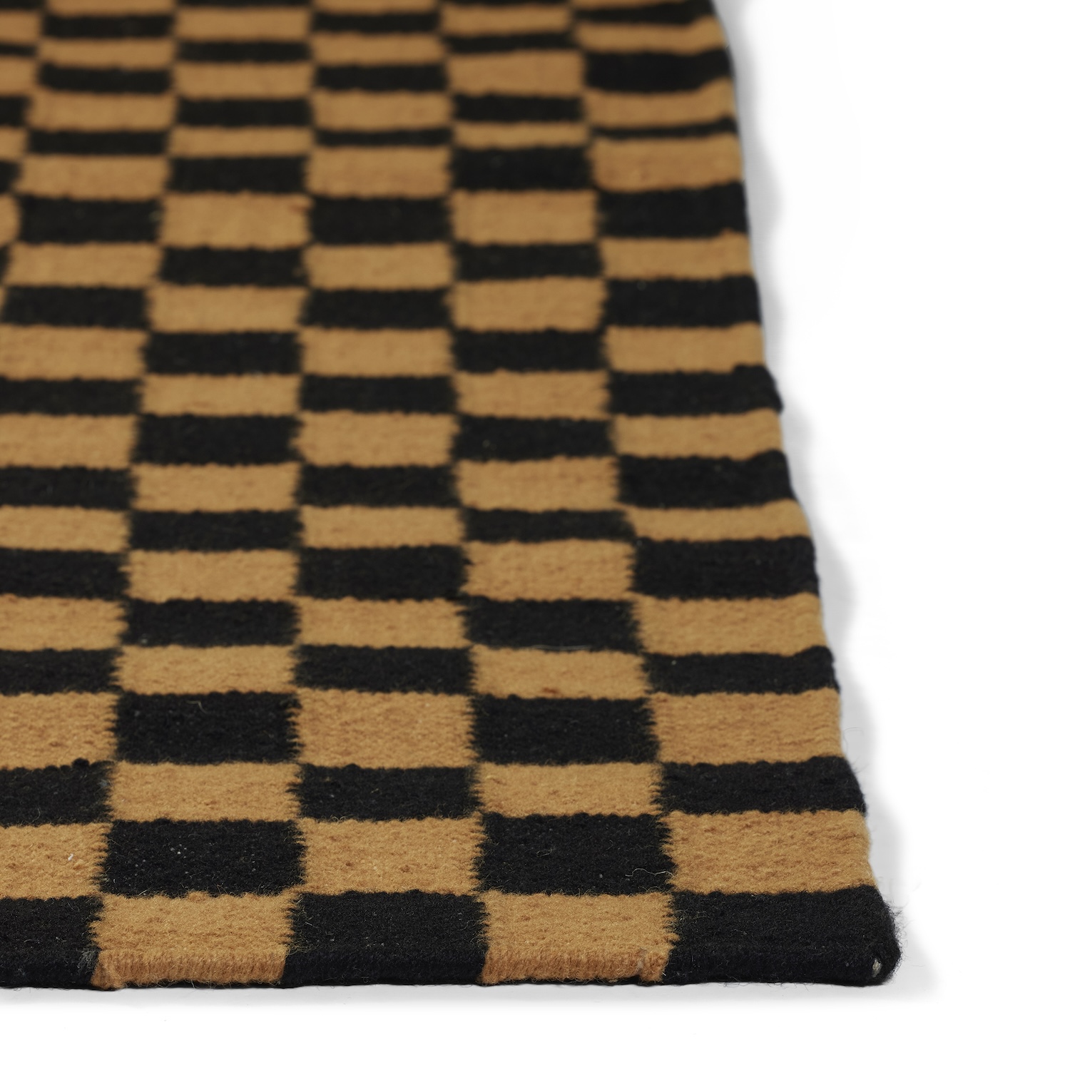 A close-up, angled shot of a rug featuring a black and tan/camel checkered pattern. The focus is on the corner and the texture of the woven material. The squares are horizontally aligned in this view. The tan/camel color is a medium, warm brown hue. The rug's edge shows a simple, woven finish, and it is photographed against a bright white background.