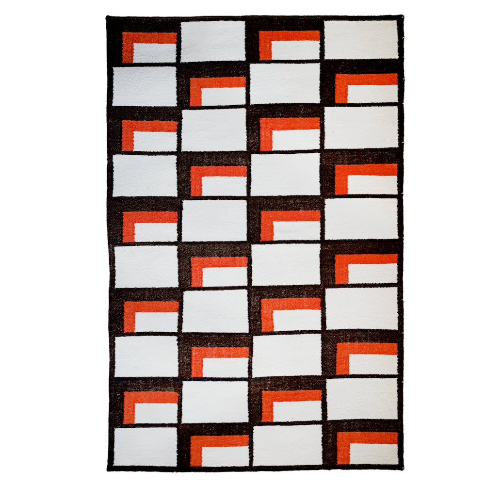 An eye-level, full shot features a hand-woven rug with a geometric pattern of white rectangles, brown rectangles, and orange L-shaped figures that outline some of the white rectangles. The rug is on a plain, gray background. The rug itself is framed by a thin black border. Each white rectangle is separated by a thin brown line. Some of the white rectangles are outlined on two sides by orange "L" shapes. The white rectangles are oriented horizontally, but the "L" shapes are on the top and left side of some of the white rectangles, while others are on the bottom and right.