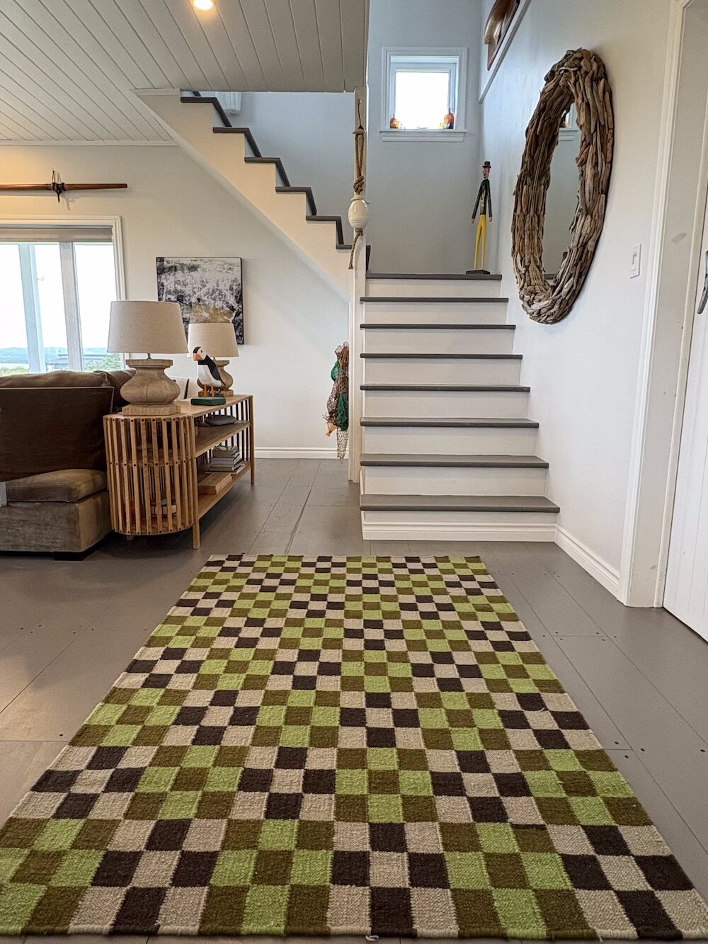 A straight-on photo shows a large entryway with a staircase leading up. A rectangular rug with a green, brown, and tan checkerboard pattern is in the middle of the floor. A couch is visible to the left, and a small table with two lamps sits in front of it. A large, circular mirror with a frame made of driftwood hangs on the wall next to the stairs. A small figure of a person stands on the second step.