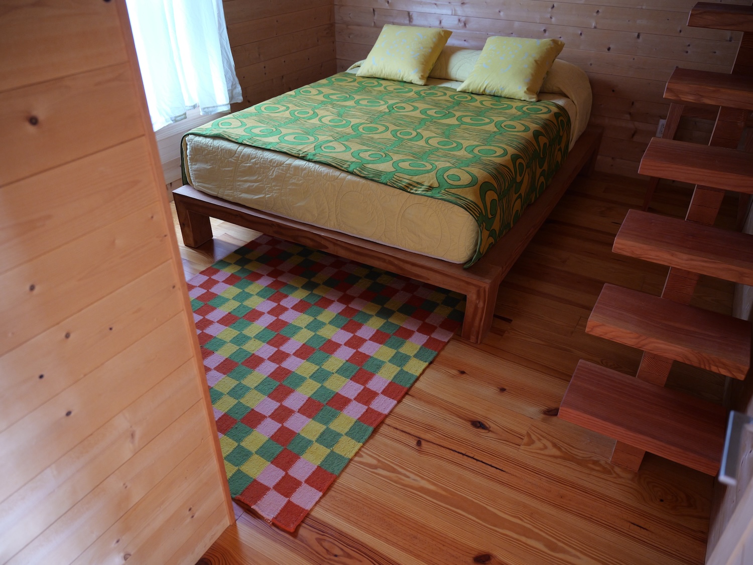 A rectangular rug with a checkerboard pattern of red, pink, yellow, and green squares is placed on a warm-toned wooden floor in a bedroom. The rug lies partially beneath a modern wooden bed frame. The bed is covered with a pale yellow comforter and a green blanket featuring a swirling geometric pattern in a darker green and yellow. Two light yellow pillows are at the head of the bed. To the left, a white curtain hangs in front of a window, letting in soft light. To the right, a set of open wooden stairs ascends, showing the underside of each step. The walls of the room are paneled with light-colored wood.