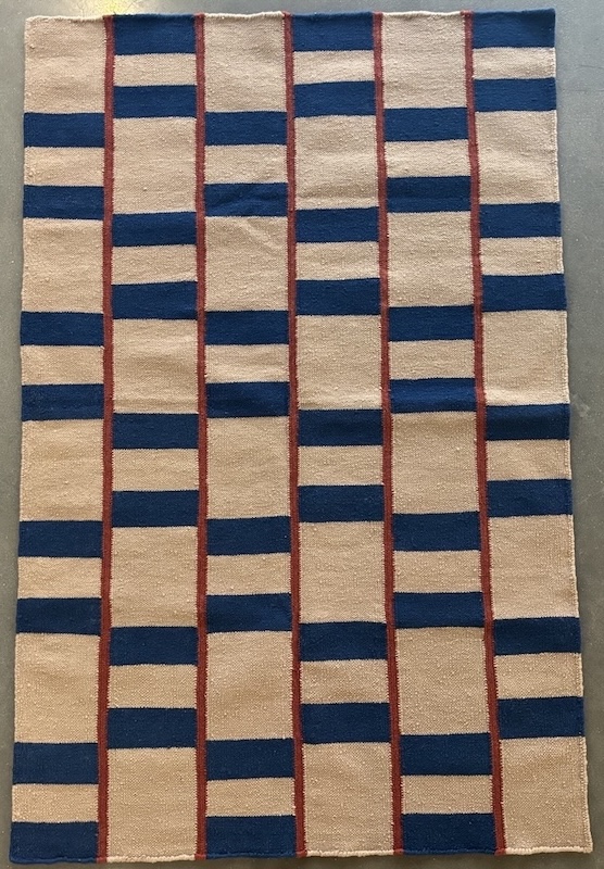 Rectangular woven rug with a beige background, navy blue horizontal stripes arranged in offset blocks, and thin vertical red stripes separating the columns.