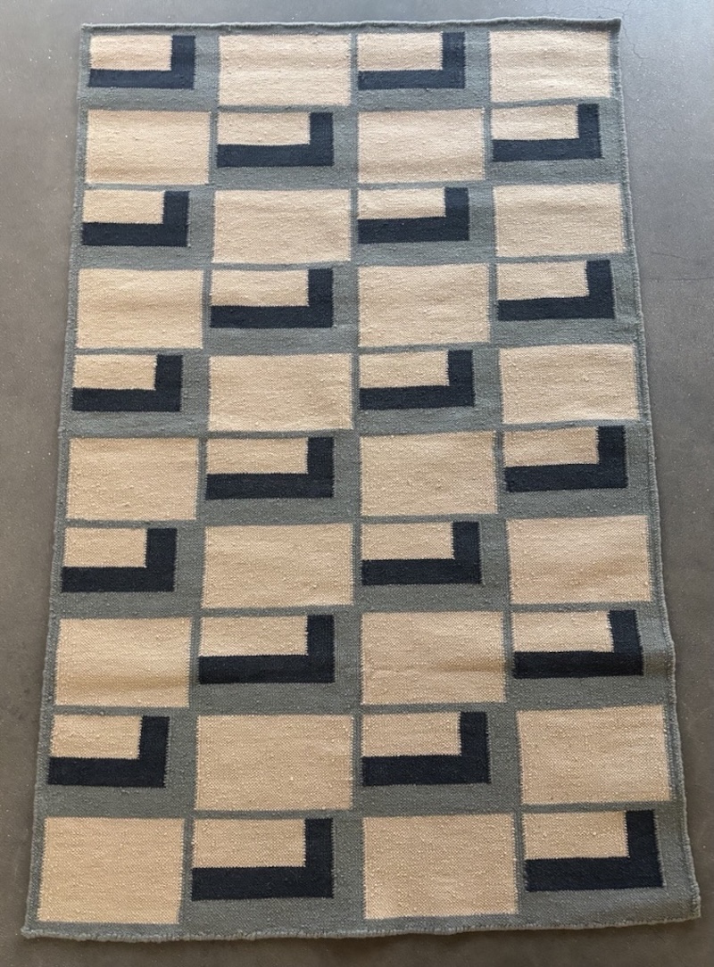 A full overhead view of a flat-woven rectangular rug on a concrete floor. The rug displays a repeating geometric pattern of cream/tan rectangular blocks arranged in a grid, divided by gray borders. Each block contains a bold navy blue L-shaped bracket in one corner, with the brackets rotating direction across the grid, creating a rhythmic, modern design.