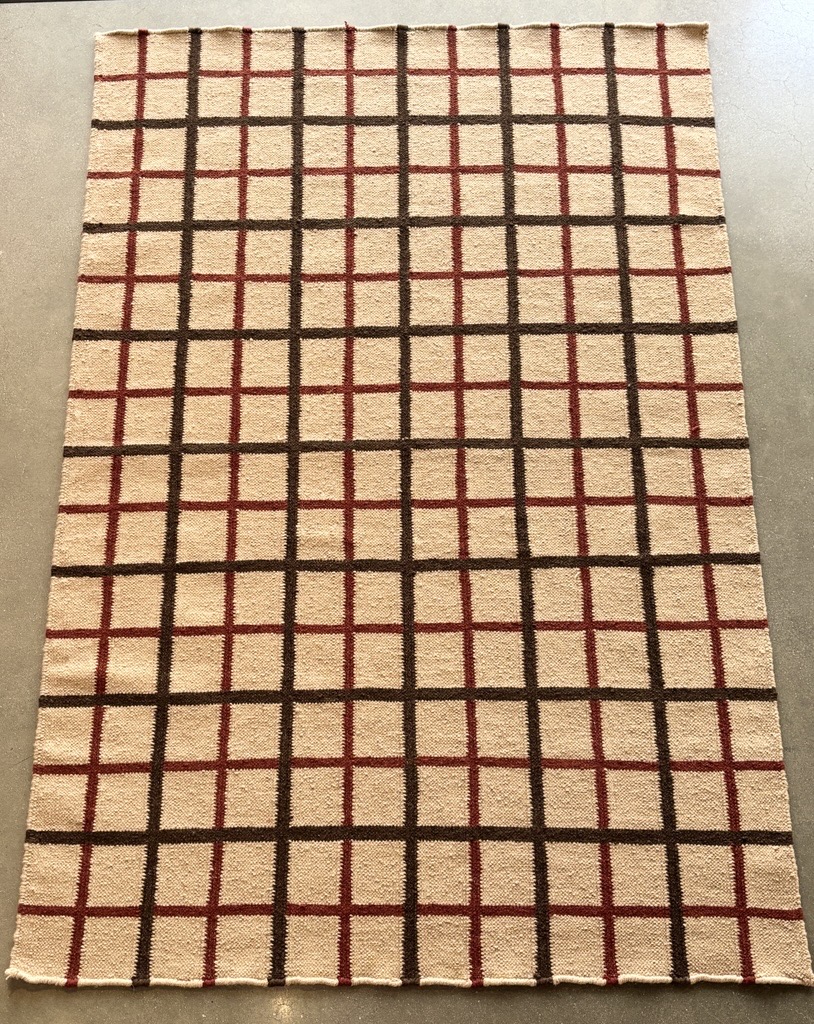 A rectangular area rug is displayed on a dark, possibly concrete or tiled, floor. The rug features a grid pattern against a light, creamy beige or off-white background. The grid is formed by intersecting lines of two distinct colors: a dark, rich brown, and a deep reddish-maroon. The dark brown lines create larger, more prominent squares, while the reddish-maroon lines subdivide these larger squares into smaller, color-accented sections within them. The rug appears to be made of a woven material with a visible texture.
