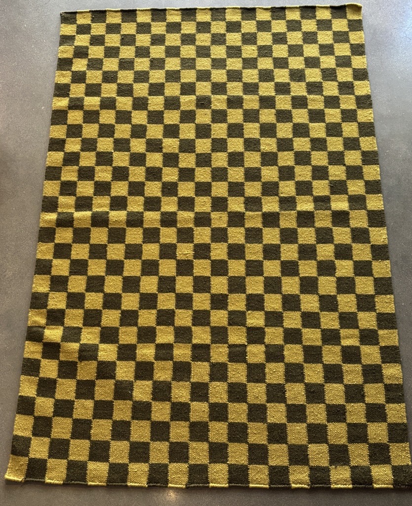 A rectangular area rug is laid out on a dark, possibly concrete or tiled, floor. The rug features a classic checkerboard pattern composed of alternating squares of two distinct colors: a muted, dark olive green or deep forest green, and a vibrant, mustard yellow or golden ochre. The squares are uniform in size and arranged in a continuous grid across the entire surface of the rug. The rug appears to be made of a woven material.