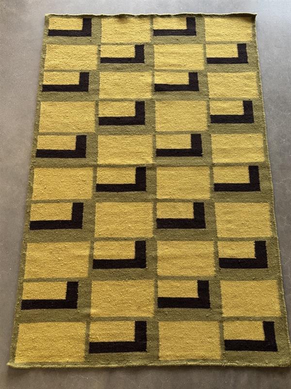 A close-up shot of a woven rug displaying a repeating geometric pattern. The primary background color is a light, muted yellow or chartreuse. Dark, almost black, L-shaped lines are arranged within and around larger, light gray-green rectangular outlines. The L-shapes are thick and solid, giving a bold visual. The woven texture of the rug is visible, showing individual threads and a slightly coarse feel.