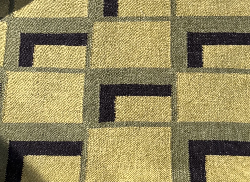 A close-up shot of a woven rug displaying a repeating geometric pattern. The primary background color of the rug is a light, muted yellow or chartreuse. Thick, dark, almost black, L-shaped lines are arranged in a repeating pattern, sometimes forming corners of implied larger squares, against a background of light gray-green rectangular outlines. The L-shapes are solid and bold, standing out against the lighter colors. The woven texture of the rug is visible, showing individual threads and a slightly coarse feel.