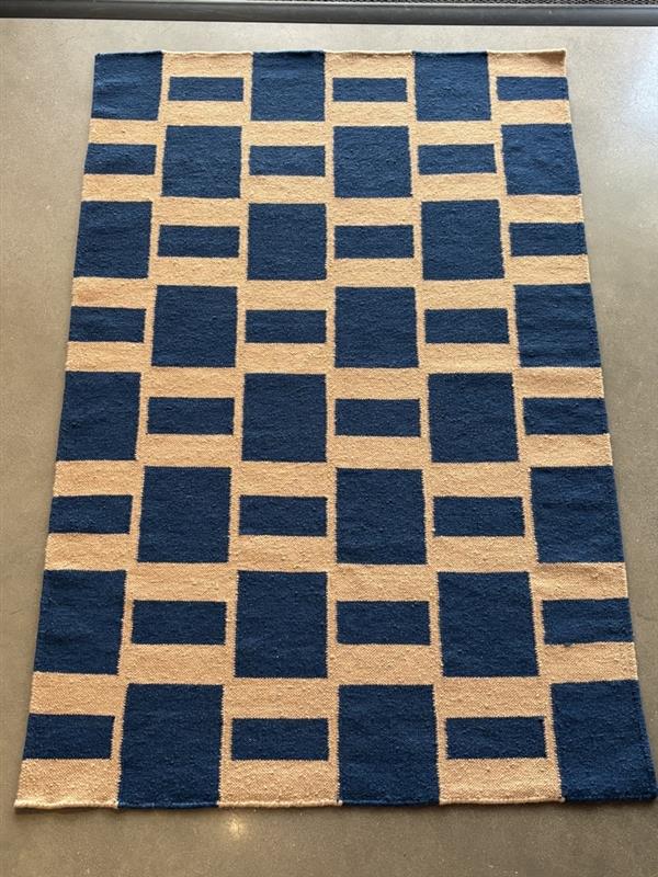 A rectangular rug with a bold, repeating geometric pattern lies on a dark gray floor. The pattern consists of solid navy blue and light beige blocks that interlock, forming a grid-like design where some beige blocks are squares and others are wider rectangles, with navy blue blocks filling the remaining spaces. The overall impression is a modern, abstract checkerboard or maze.