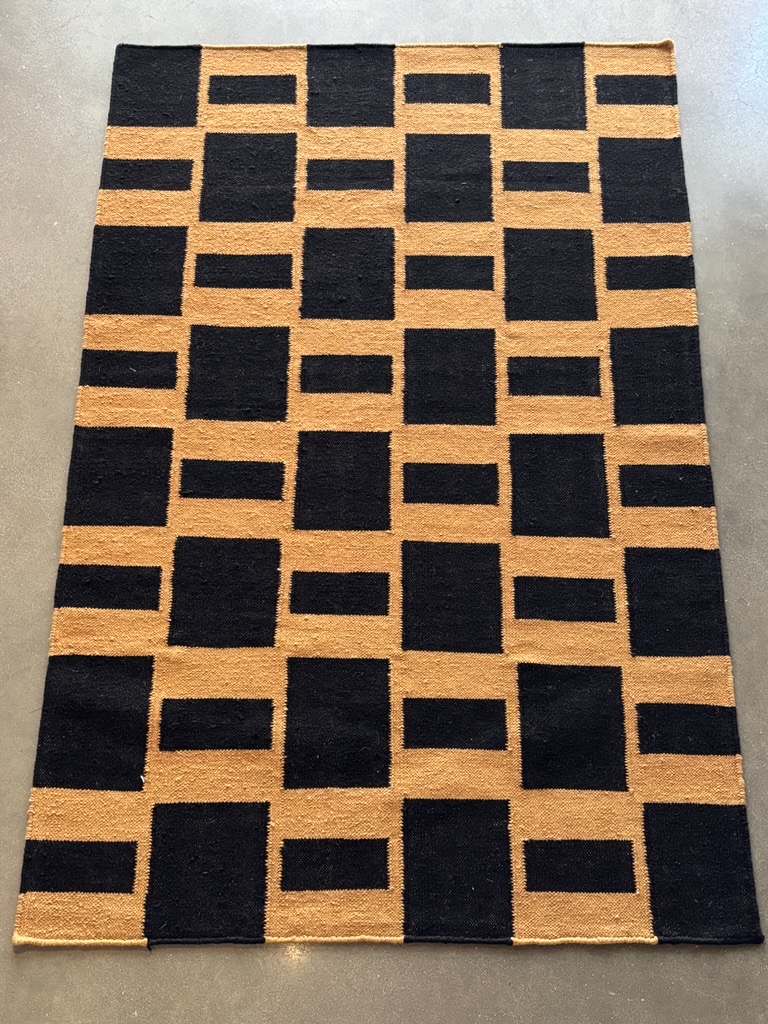 A rectangular rug featuring a bold geometric pattern of interlocking solid black and solid dark tan blocks is laid out on a dark gray floor. The tan blocks form both squares and horizontal rectangles, while the black blocks fill the remaining spaces, creating a structured, maze-like design.