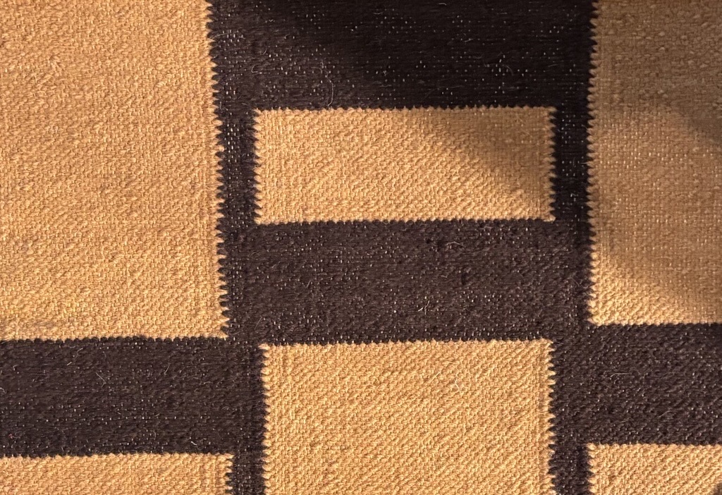 A close-up view of a woven rug shows a section of its geometric pattern. The pattern features alternating solid blocks of dark brown and golden tan. The tan blocks are seen as both a large square and a smaller horizontal rectangle, while the dark brown forms a wide horizontal band and a vertical section, all fitting together. The texture of the tightly woven fibers is clearly visible, with slight variations in color intensity within the tan areas.
