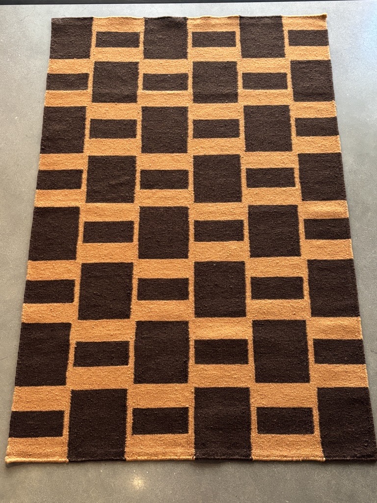 A rectangular rug with a striking geometric pattern lies flat on a dark gray concrete floor. The pattern is composed of interlocking blocks in two colors: a rich, earthy brown and a warm, golden tan. The tan blocks form both squares and wider horizontal rectangles, while the brown blocks fill the remaining spaces, creating a structured, maze-like design. The edges of the rug appear neatly finished.