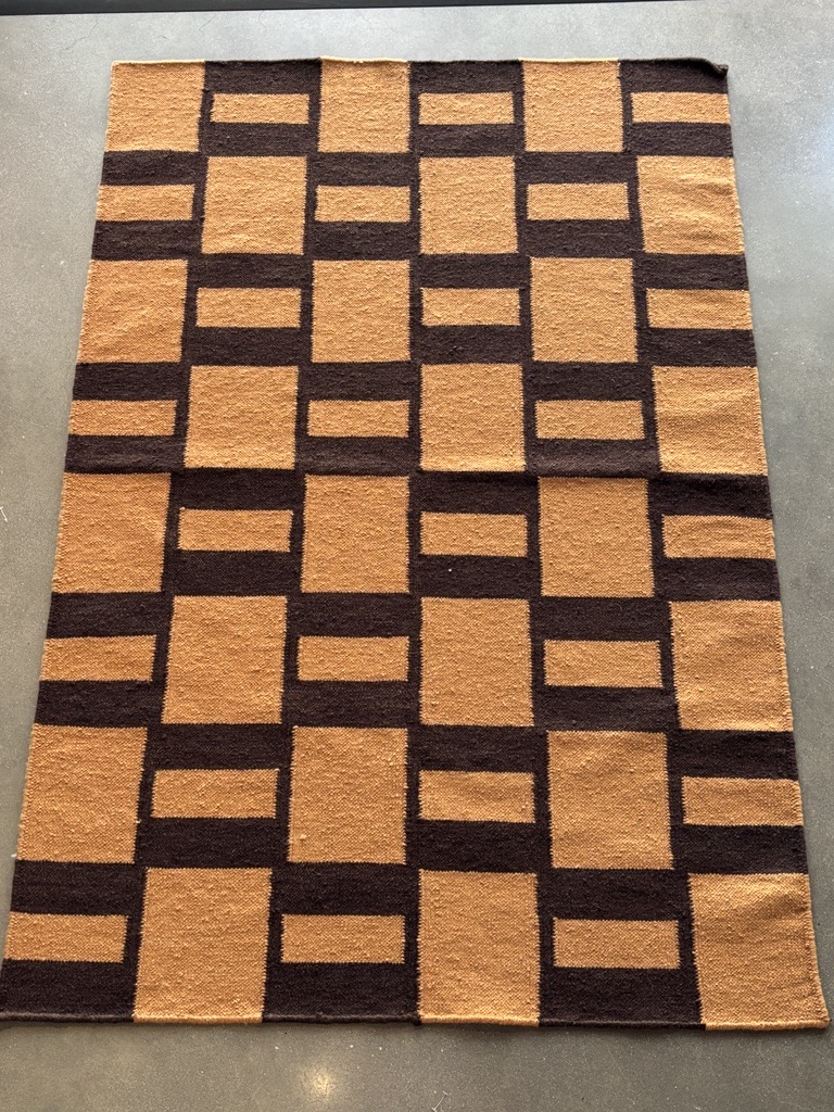 A rectangular rug with a striking geometric pattern lies flat on a dark gray concrete floor. The pattern is composed of interlocking blocks in two colors: a rich, earthy brown and a warm, golden tan. The golden tan blocks form both squares and wider horizontal rectangles, while the brown blocks fill the remaining spaces, creating a structured, maze-like design. The edges of the rug appear neatly finished.