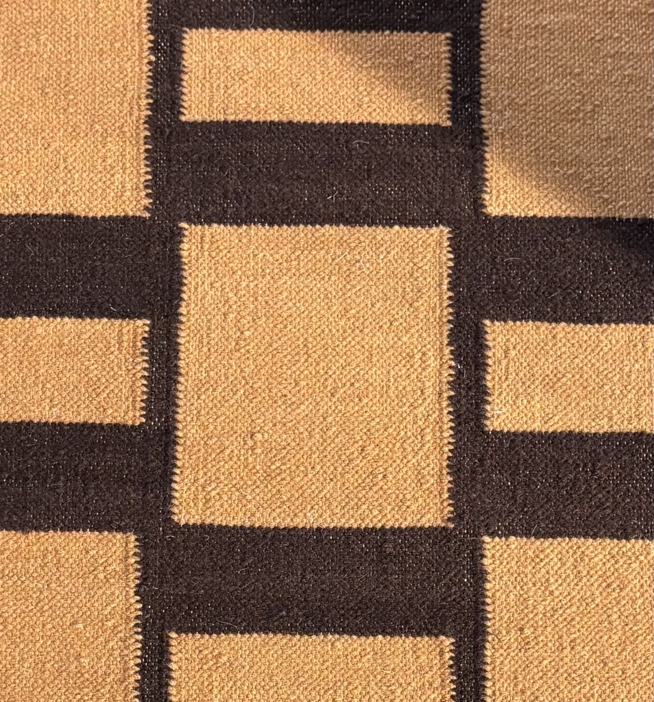 A close-up overhead shot of a woven rug's surface, showing a section of its geometric pattern. The pattern is created by interlocking blocks of dark, rich brown and a warm, golden tan. The golden tan appears in both square and horizontal rectangular shapes, while the dark brown fills the spaces between them, forming a grid. The texture of the woven fibers is clearly visible, with a slight raised quality to the individual strands.