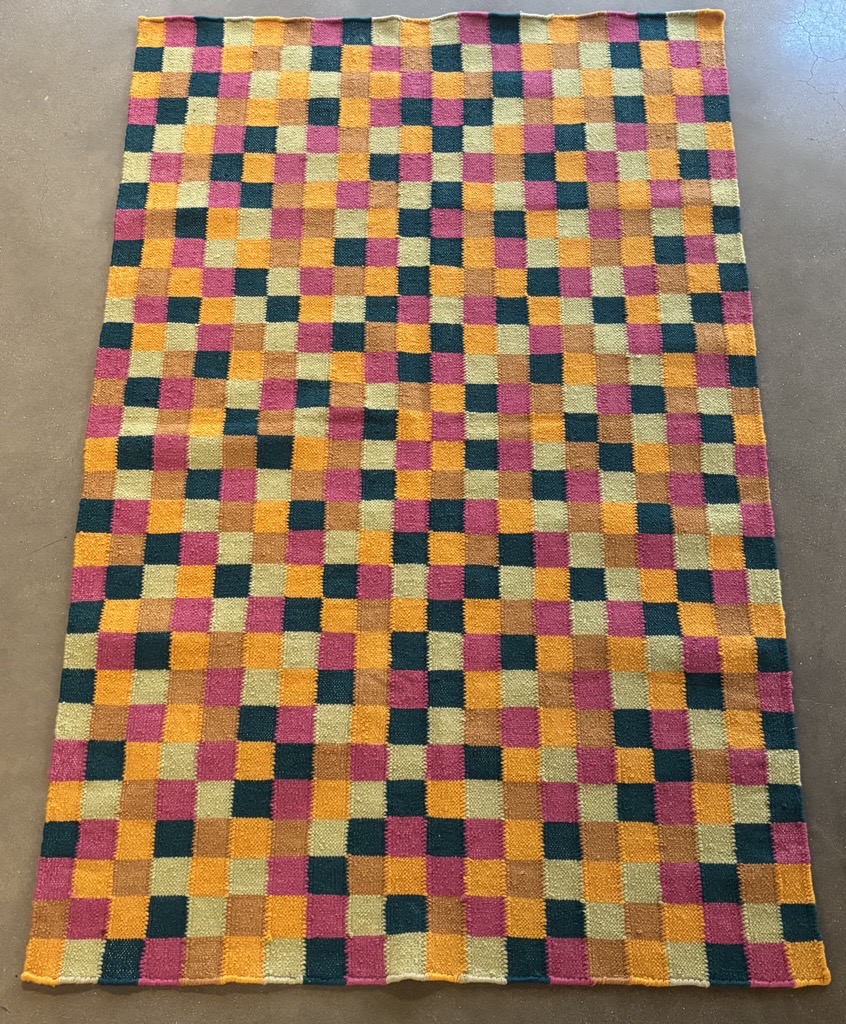 A full view of the same textile laid flat on a smooth concrete floor. The entire surface is covered in a repeating checkerboard pattern with the same five colors: magenta, orange, teal blue, beige-gray, and brown. The textile is rectangular, and the pattern is evenly distributed across it. The edges are mostly straight with a slightly undulating appearance, suggesting it is handmade. The overall effect is bold, colorful, and visually rhythmic.
