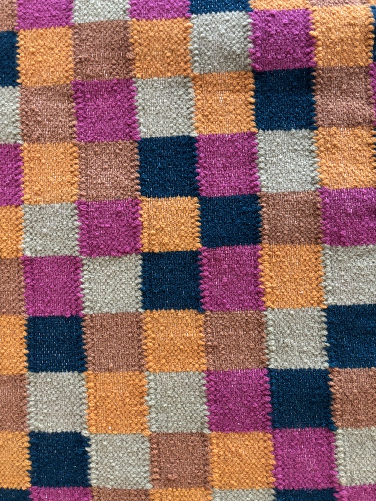 A close-up view of a handwoven textile featuring a checkerboard pattern made up of small squares. The squares are arranged in alternating colors: bright magenta, burnt orange, deep teal blue, beige-gray, and medium brown. The texture of the woven yarn is clearly visible, with each square appearing slightly raised and distinct. The tight weave and high contrast between the colors create a vibrant, tactile surface.