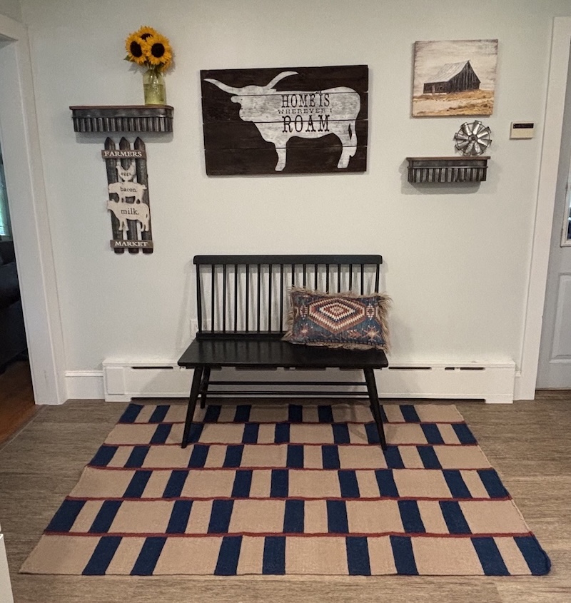 Farmhouse-style entryway with a black wooden bench on the beige rug with navy and red striped pattern, wall decor including a longhorn silhouette sign reading “Home is wherever I roam,” small shelves, a barn picture, and sunflowers in a vase.