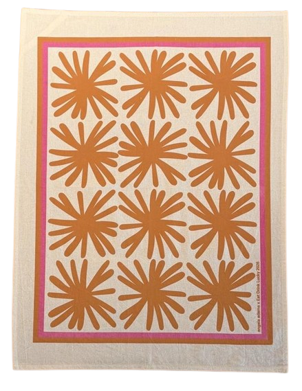 A rectangular tea towel with a natural linen background featuring a grid of twelve bold, hand-drawn orange daisy-like burst motifs arranged in four rows of three. The design is framed by a hot pink border. Small printed text appears in the bottom right corner. The overall aesthetic is graphic, playful, and mid-century modern.