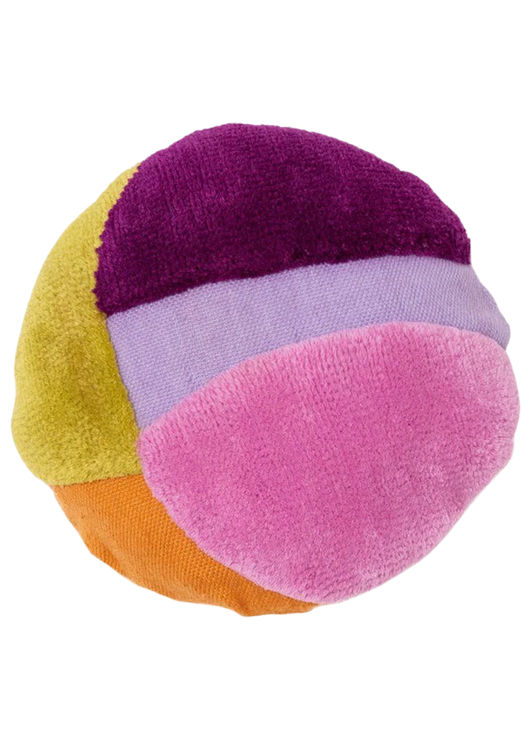 A soft, round decorative pillow composed of four curved, wedge-shaped panels sewn together. Each panel is a different color and appears to have a plush or terry-cloth texture. The colors are: deep purple (top), light lavender (second from top), bright pink (middle right, largest visible section), mustard yellow-green (left), and a sliver of orange (bottom). The pillow is shown against a transparent or white background. It resembles a small, colorful, oversized ball or beach ball shape.