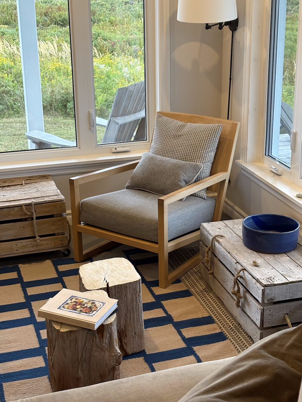 A photo shows a corner of a living room with a large window and a light-colored armchair with gray cushions and pillows. A small, square rug with a grid of navy blue rectangles on a beige background is partially visible on the floor. In front of the chair are two small tables made of unvarnished tree stumps. A book with a colorful cover is on the taller stump. To the left of the chair is a wooden crate, and to the right of the chair is another wooden crate with a dark blue bowl on top. A glimpse of a deck with an Adirondack chair is visible through the window.