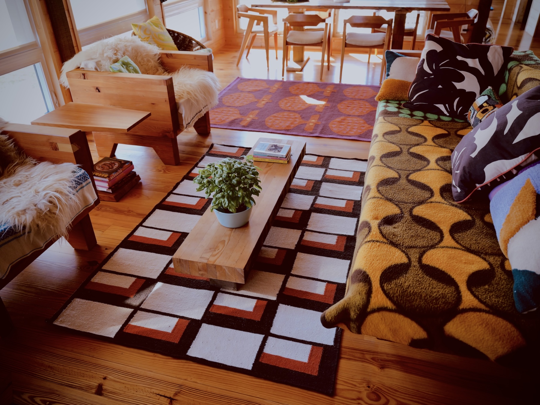 A warm and inviting living room with wooden walls and floors is shown in daylight. The centerpiece is a rectangular flatwoven rug featuring a bold, geometric pattern made up of black rectangles and right angles interlocked with olive green borders. The pattern is arranged in a grid across the entire rug. The rug has a textured appearance and sits on a flat surface, photographed from above with no surrounding furniture visible in the image. The overall aesthetic is modern and minimal, with strong visual contrast between the dark and muted colors.