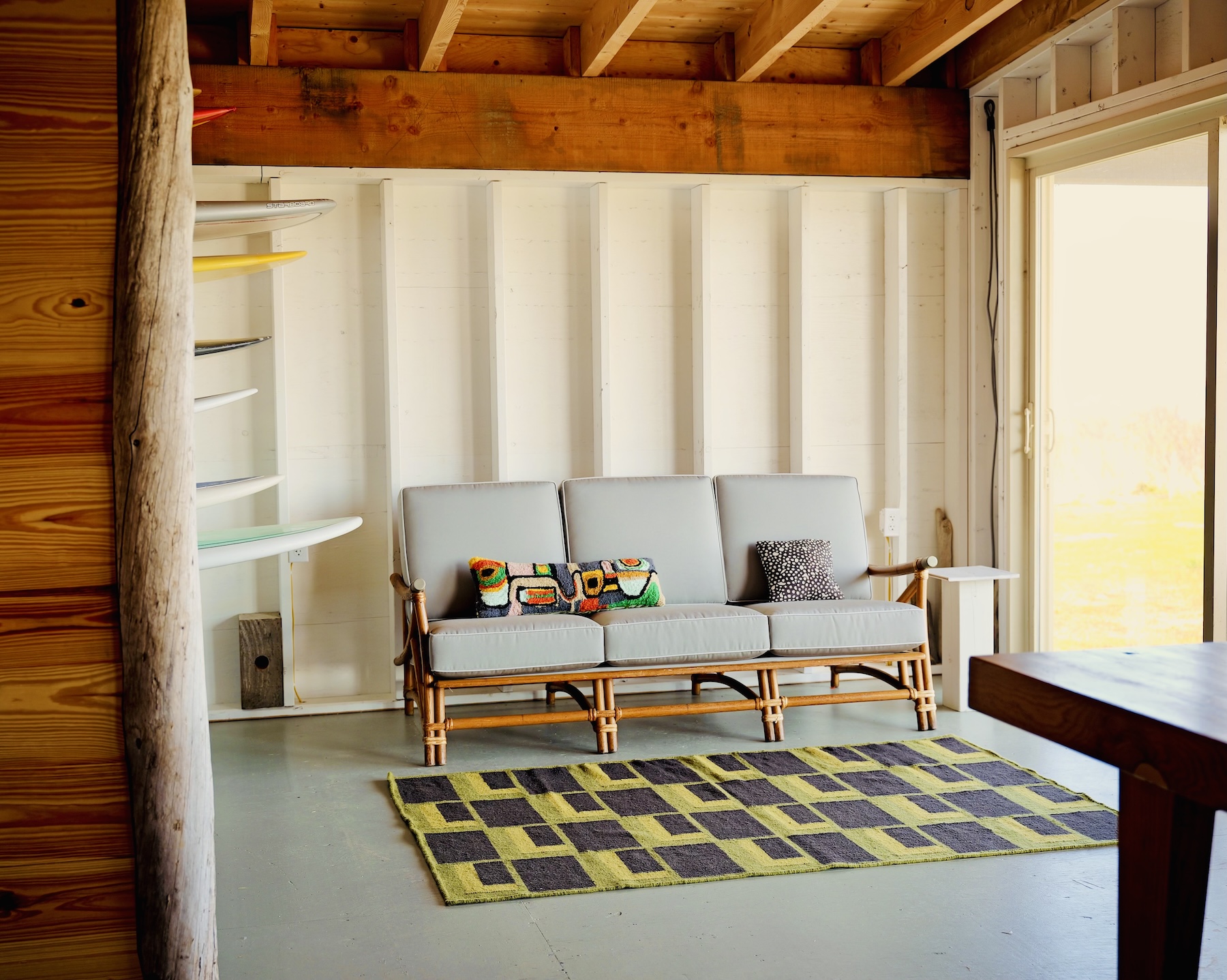 A bright, cozy room with an exposed wood beam ceiling and white paneled walls features a modern coastal aesthetic. Centered along the back wall is a light gray cushioned rattan sofa with three seats and wooden arms, adorned with two decorative throw pillows—one multicolored with abstract shapes and the other black with white dots. In front of the sofa lies a rectangular area rug with a geometric pattern of interlocking dark charcoal and olive green rectangles. To the left of the sofa, multiple surfboards are stored vertically on wall-mounted racks, adding a playful, beachy vibe. Natural light pours in through a large glass sliding door on the right, illuminating the space with a warm, inviting glow. The floor is painted in a soft, muted green.