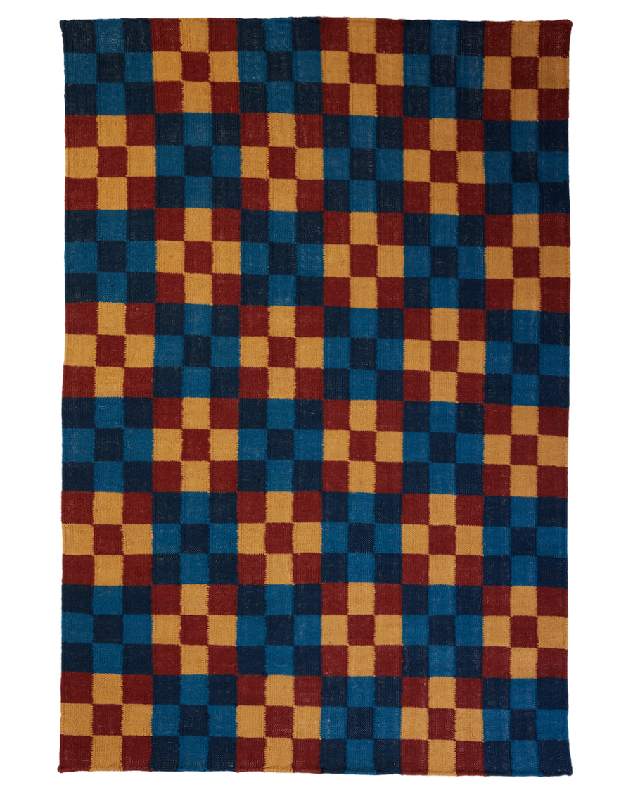 A rectangular, hand-woven rug with a checkerboard pattern. The colors alternate between deep teal blue, mustard yellow, and a muted, dark cranberry red. The rug is shot from a top-down, straight-on perspective against a white background, clearly showing its full outline and texture. The edges are unfinished or fringeless. The pattern consists of rows and columns of approximately square blocks of color.